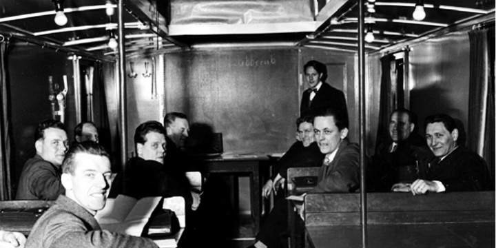 Svartvit bild på studerande som sitter i bänkrader i den klassrumsliknande tågvagnen.